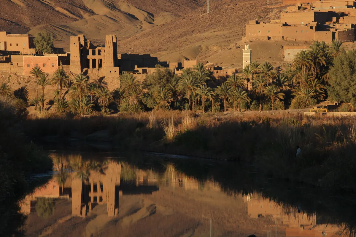 Une kasbah en terre, des palmiers et un minaret se reflètent dans le Drâa à Agdz, dans une lumière dorée