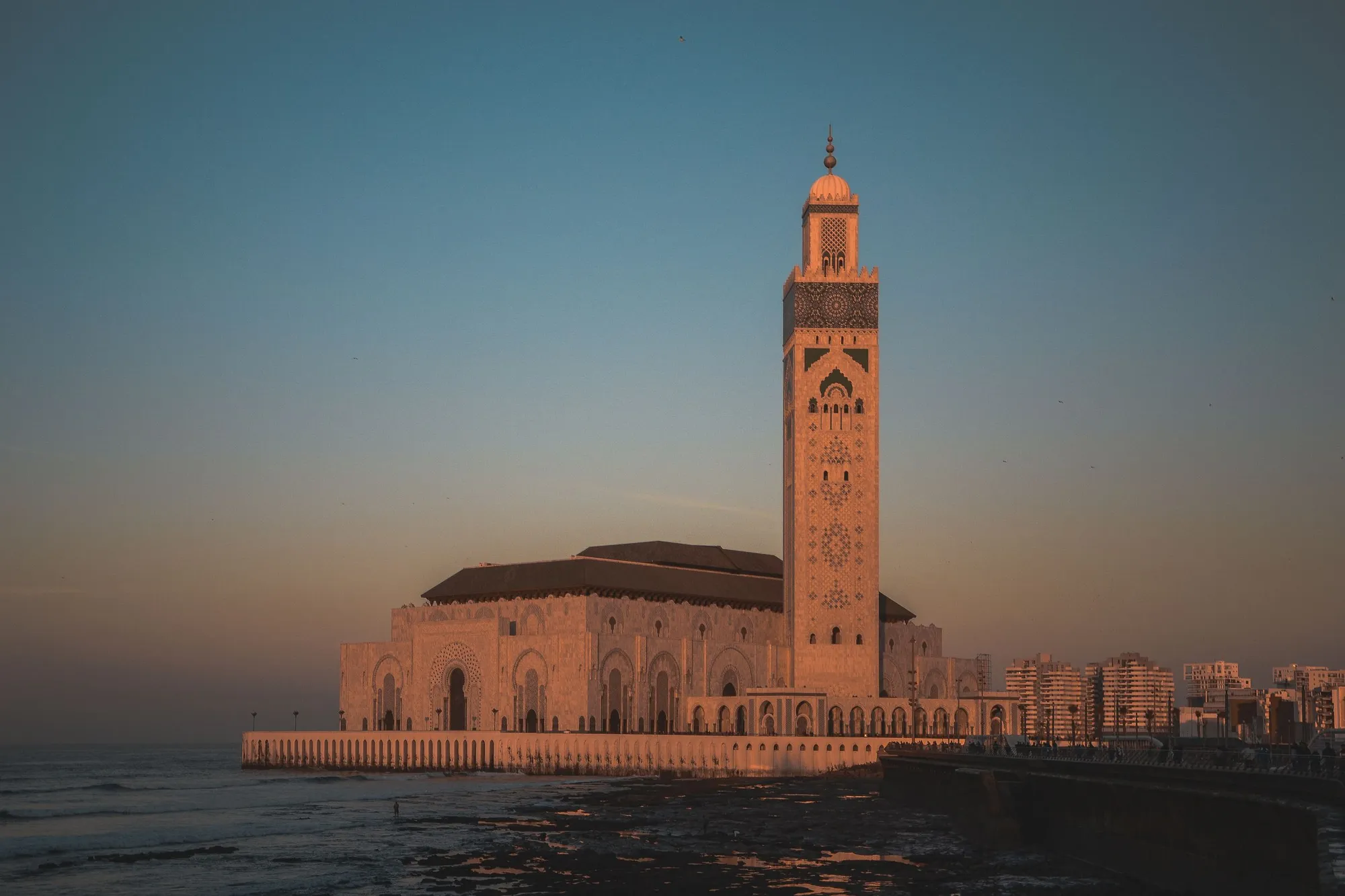 The Hassan II Mosque and its minaret at sunset, standing on the edge of the Atlantic with the city behind