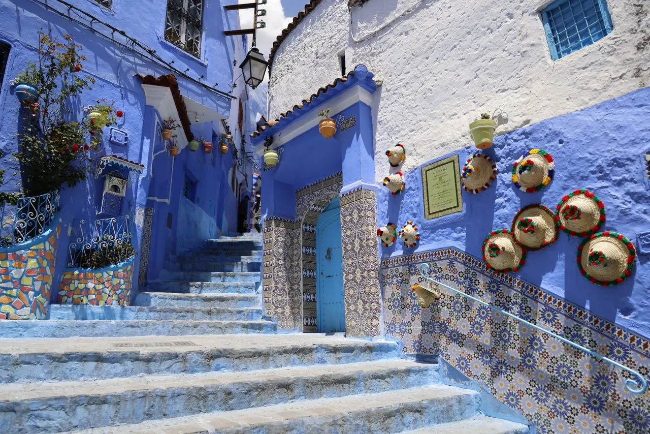 A blue stairway in Chefchaouen, lined with zellige tiles and straw hats with pompoms hung on the wall
