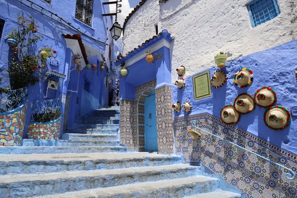 A blue stairway in Chefchaouen, lined with zellige tiles and straw hats with pompoms hung on the wall