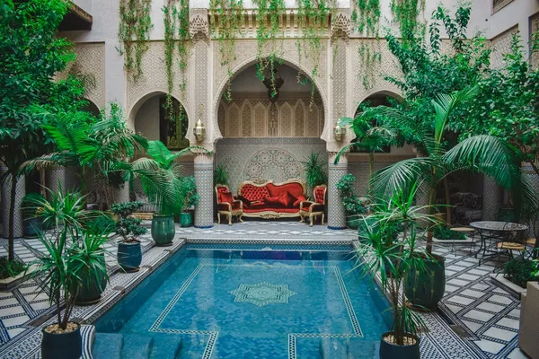 The courtyard of a riad with its zellige pool, palm trees and a red sofa under an arch