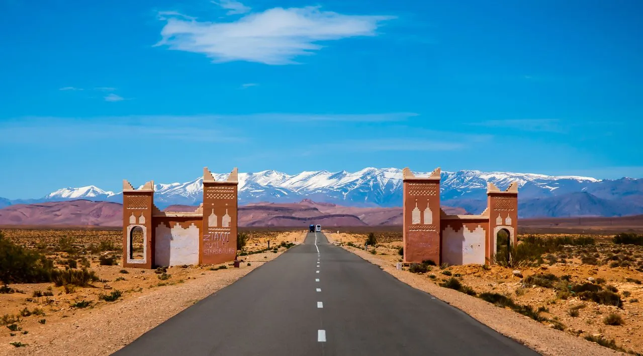 A road passing under a gate built like a kasbah, with the snowy Atlas on the horizon