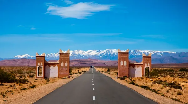 A road passing under a gate built like a kasbah, with the snowy Atlas on the horizon