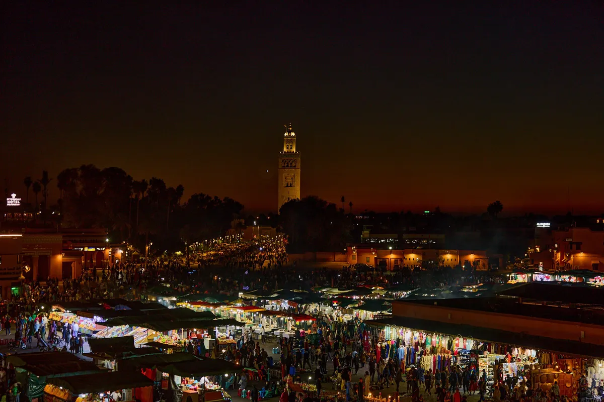 Jemaa el Fna square crowded with lit stalls at dusk, the Koutoubia minaret standing against the evening sky