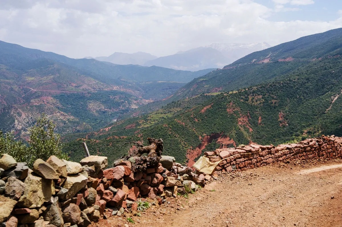A dirt track lined with a dry stone wall, overlooking green mountain valleys with higher peaks in the distance