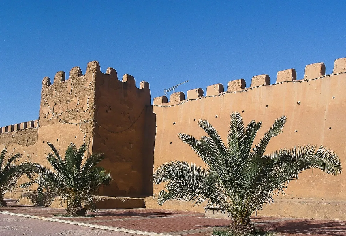 Les remparts crénelés en terre ocre de Taroudant, avec une tour carrée et de jeunes palmiers au pied du mur