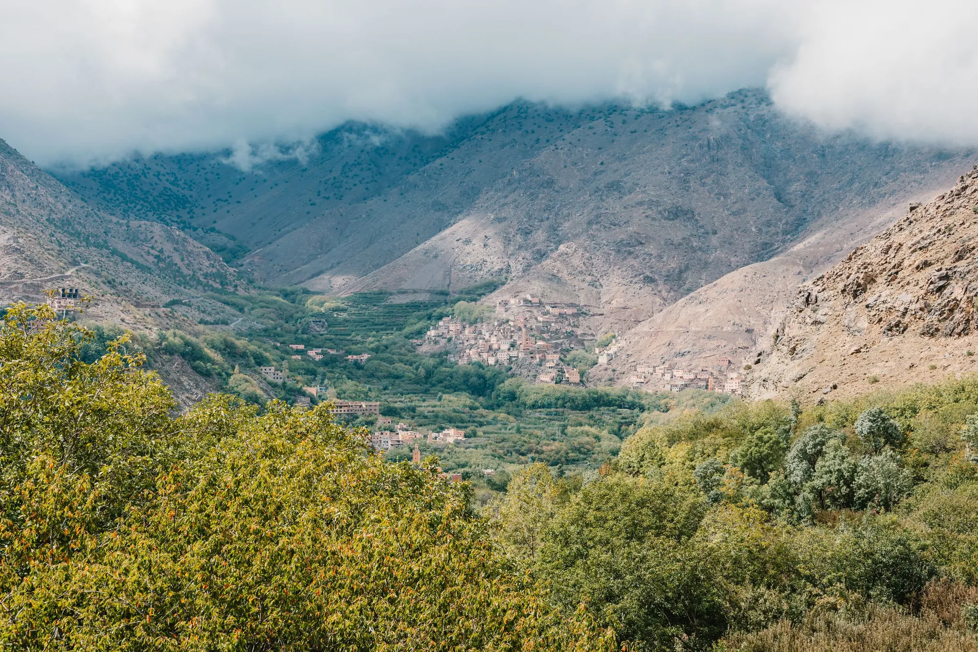 A village of earth houses in a green valley of the Toubkal massif, below mountains capped with cloud