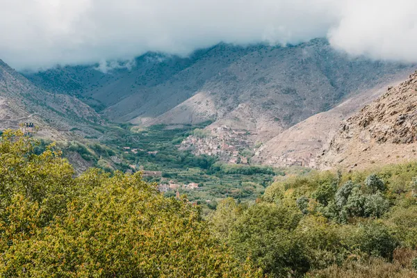 A village of earth houses in a green valley of the Toubkal massif, below mountains capped with cloud