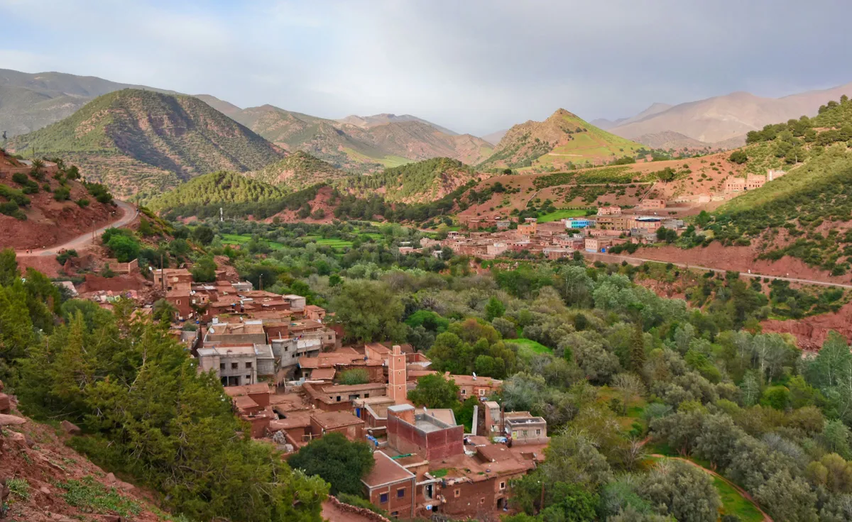 Red earth houses of a mountain village among trees, with green hills and a winding road behind
