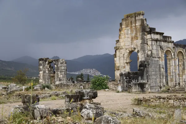 The stone arches of the Roman basilica of Volubilis under a stormy sky, with the white town of Moulay Idriss on the hill behind