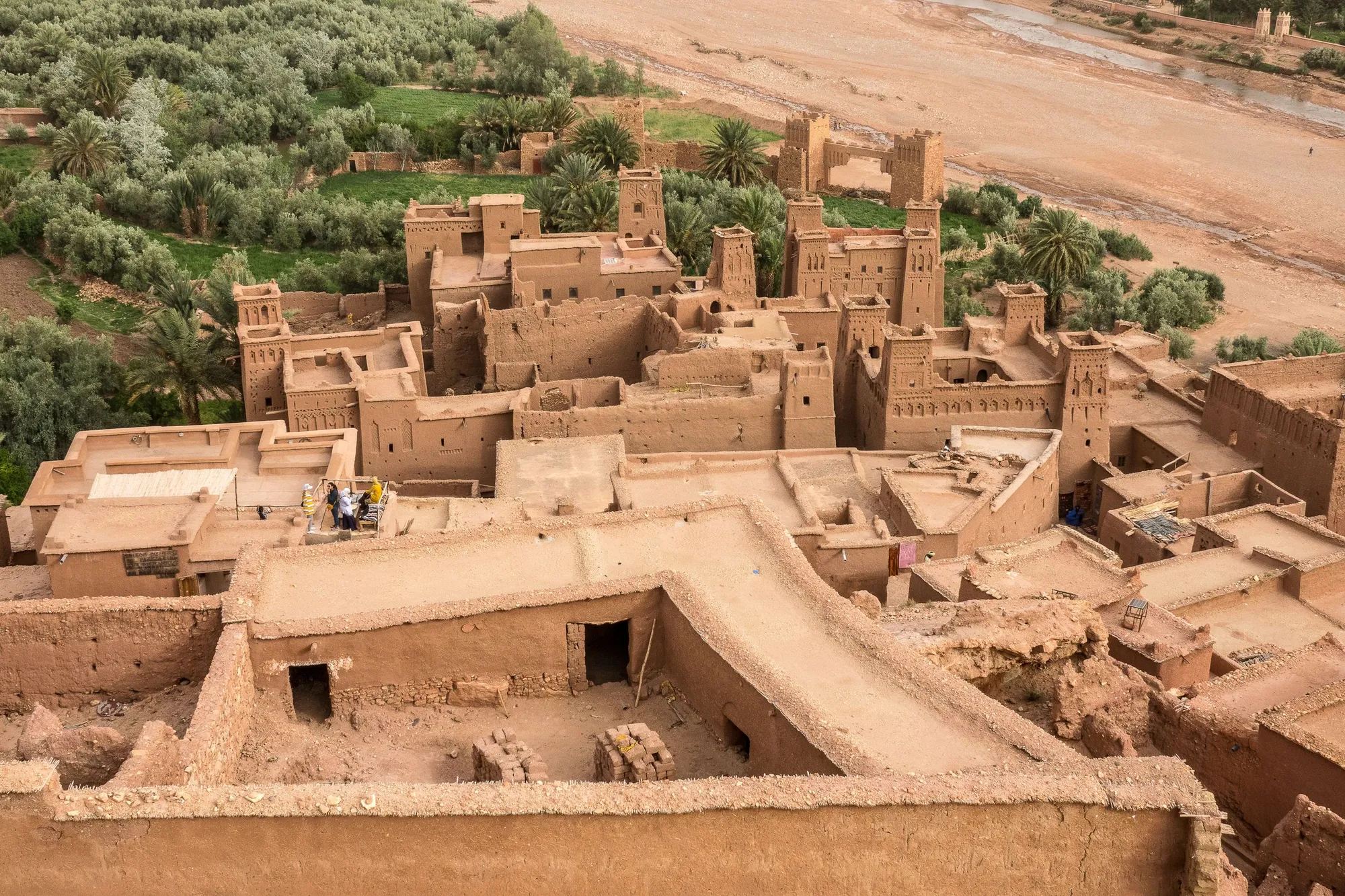 The earthen towers and terraces of the ksar of Ait Benhaddou seen from above, with palm trees and a green valley behind them