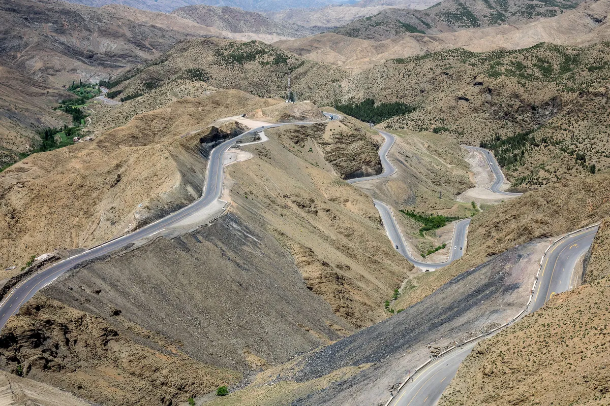 A road climbing in long hairpin bends across dry, rocky mountains, with patches of green in the folds of the valley