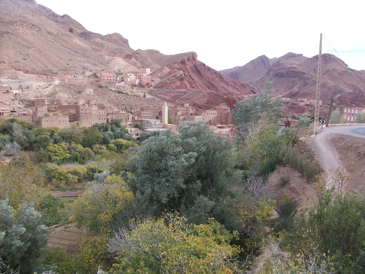 A village of earthen houses on a slope of red mountains, above a valley full of green and yellow trees, with a road curving along the hillside